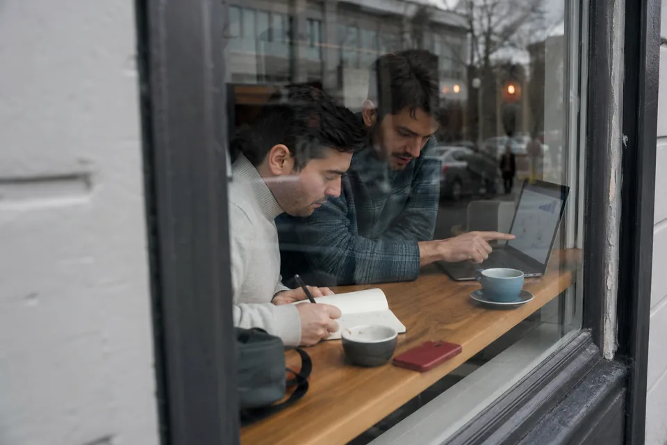 Two co-founders at a cafe window table adding up broker costs on a notepad