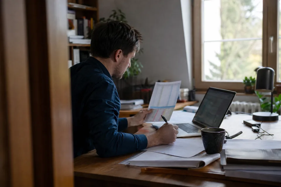 Founder at a home office desk comparing a domain asking price with sold prices on a laptop and a printout